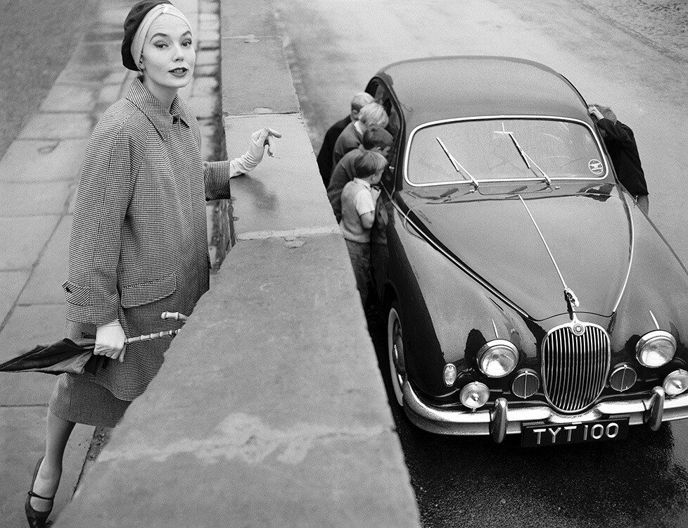 Пэт Годдард стоит у Jaguar 3.4 в черно-белом клетчатом пальто от Aquascutum и коричневой замшевой шляпе от Dolores Boutique. Vogue, ноябрь 1957 года. Фотограф Норман Паркинсон