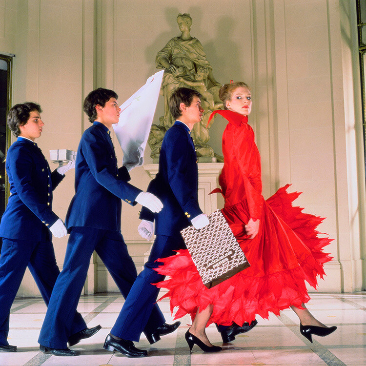 Андреа Холтерхоф в пышном красном вечернем платье, за ней следуют три посыльных в холле отеля Meurice в Париже, Франция, Vogue 1979. Фотограф Норман Паркинсон