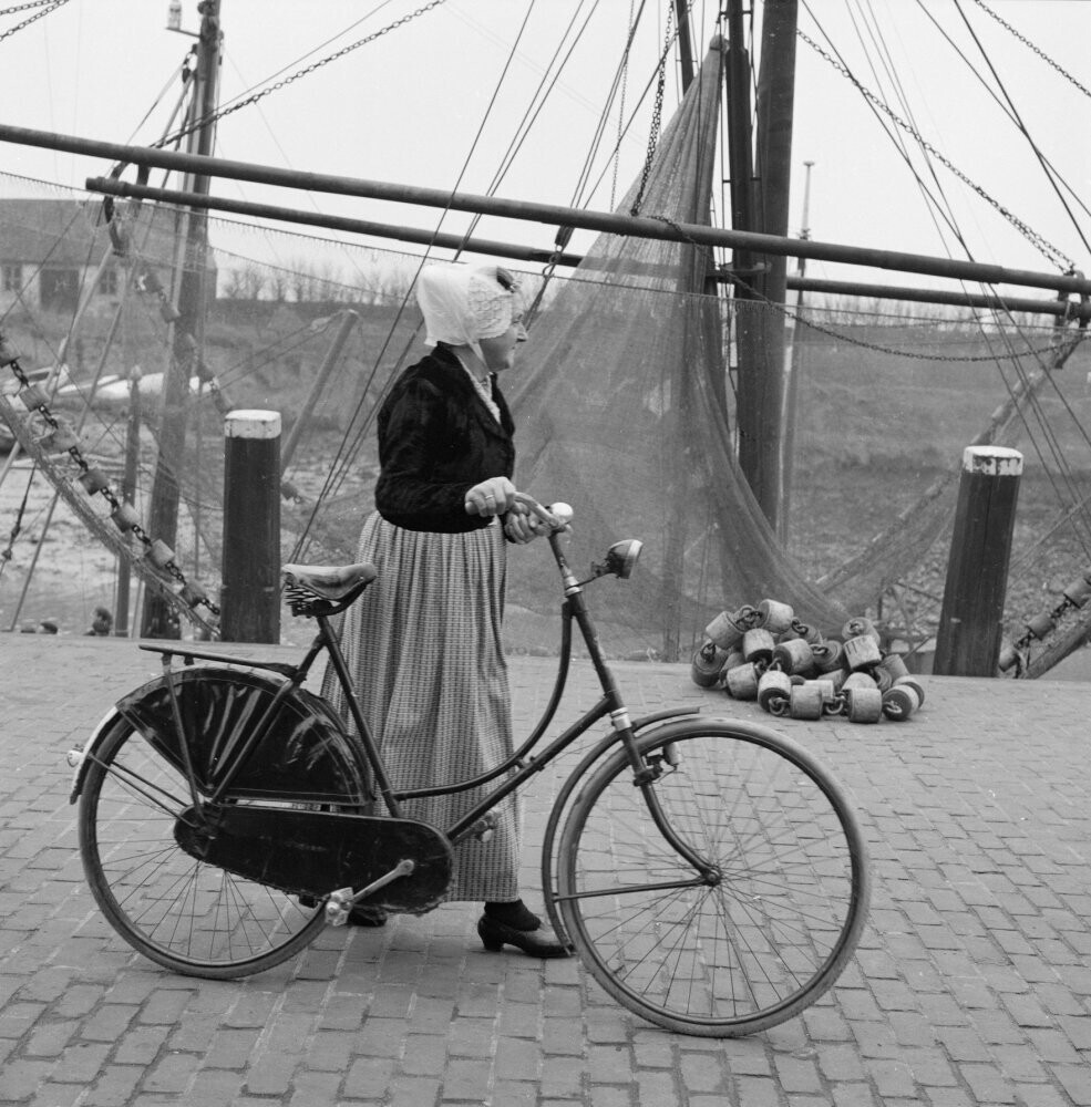 Женщина в традиционном костюме с велосипедом в рыбацком порту, 1961 год ...