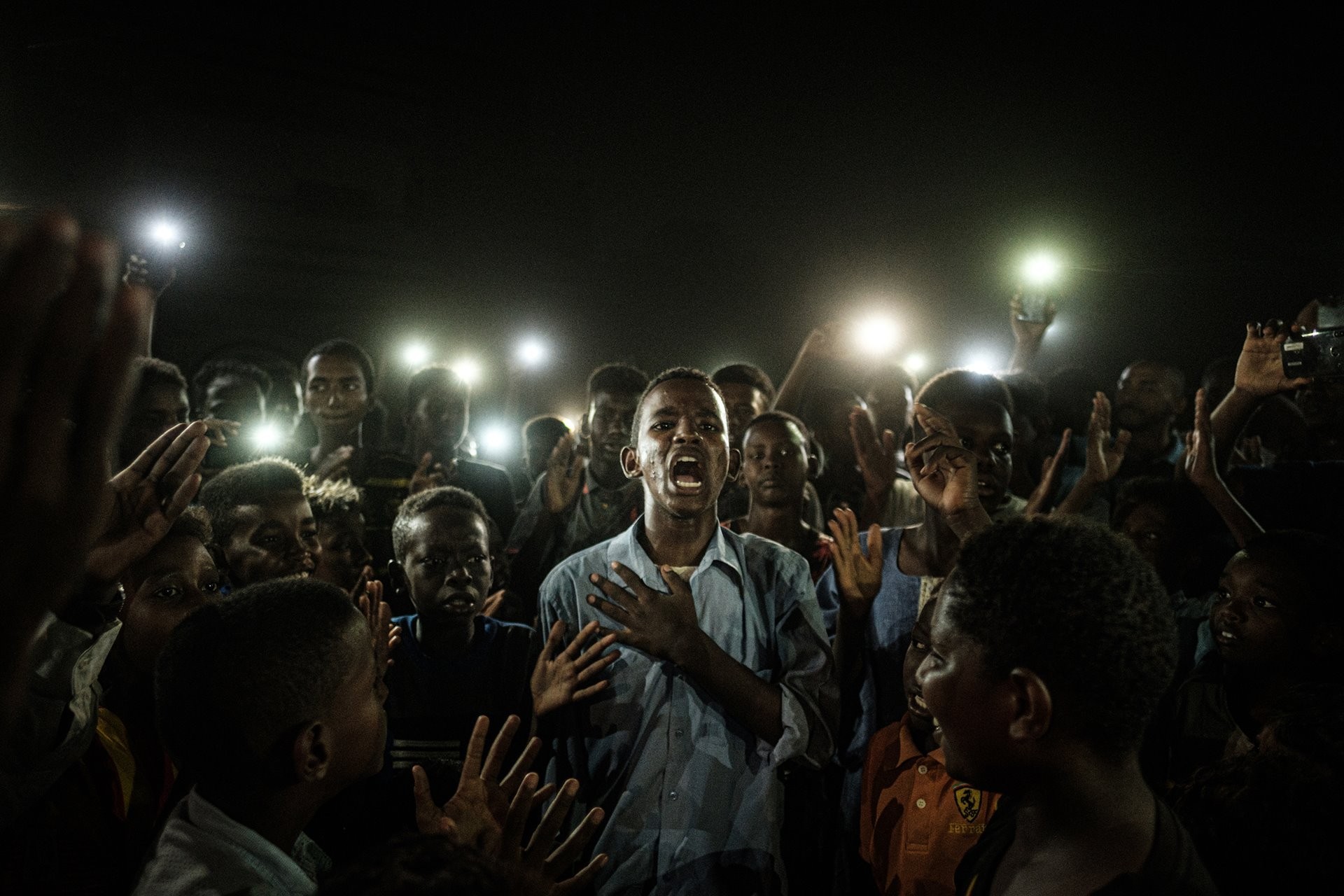 Фото года, 2020. Участник протестов против режима Омара аль-Башира читает стихи в свете телефонов при отключенной электроэнергии в Хартуме, Судан. Автор Ясуёси Тиба
