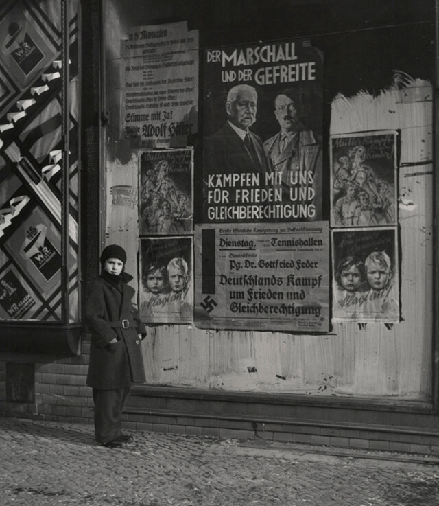 Дочь Вишняка Мара позирует перед предвыборным плакатом Гинденбурга и Гитлера с надписью «Маршал и капрал сражайтесь с нами за мир и равные права», Вильмерсдорф, Берлин, 1933 г. Фотограф Роман Вишняк