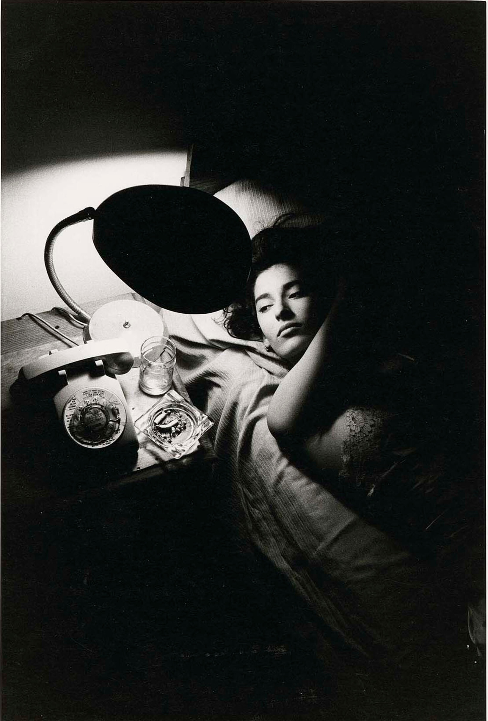 Женщина лежит на кровати рядом с тумбочкой с телефоном, лампой и пепельницей, 1986 г. Фотограф Дэвид Сайднер